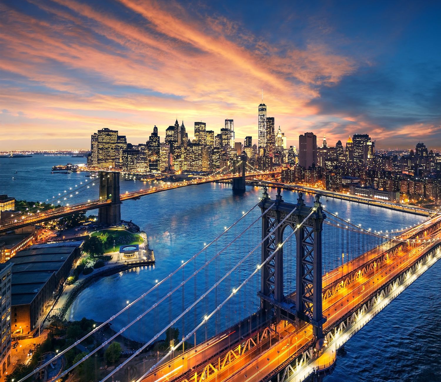 Aerial view of New York City skyline at sunset with lit bridges and skyscrapers reflecting in the river.