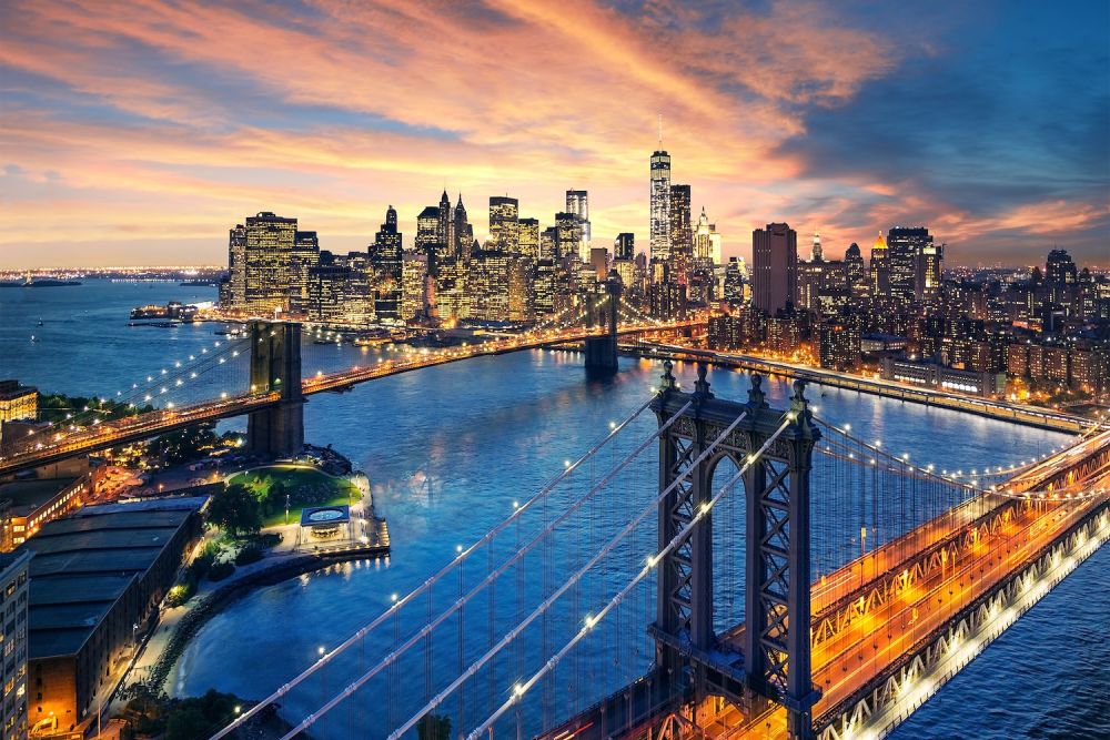 Aerial view of New York City skyline at sunset with lit bridges and skyscrapers reflecting in the river.
